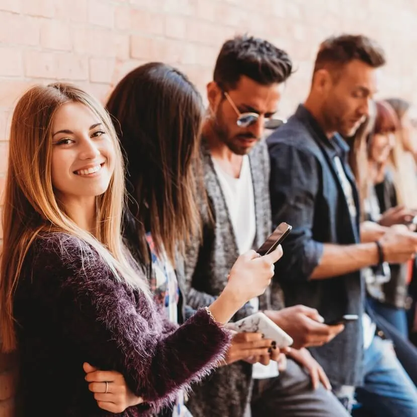 A group of young adults stands against a brick wall, looking at their smartphones. One woman in the foreground smiles directly at the camera while the others use Online SMS on their devices.
