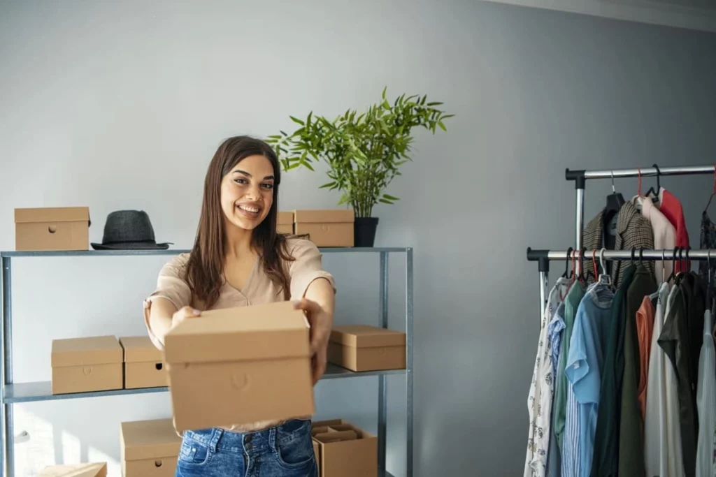 Eine lächelnde Frau steht in einem Raum und hält einen Karton in Richtung der Kamera, bereit für Abholungseinladungen per WhatsApp. Hinter ihr stehen Regale mit Kartons, ein Hut, eine Topfpflanze und ein Kleiderständer mit verschiedenen Kleidern.