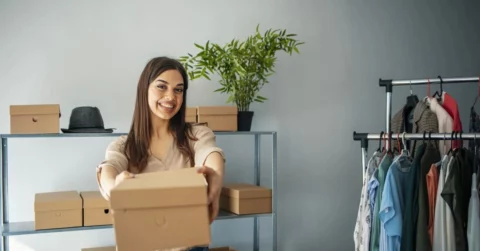 Eine lächelnde Frau steht in einem Raum und hält einen Karton in Richtung der Kamera, bereit für Abholungseinladungen per WhatsApp. Hinter ihr stehen Regale mit Kartons, ein Hut, eine Topfpflanze und ein Kleiderständer mit verschiedenen Kleidern.