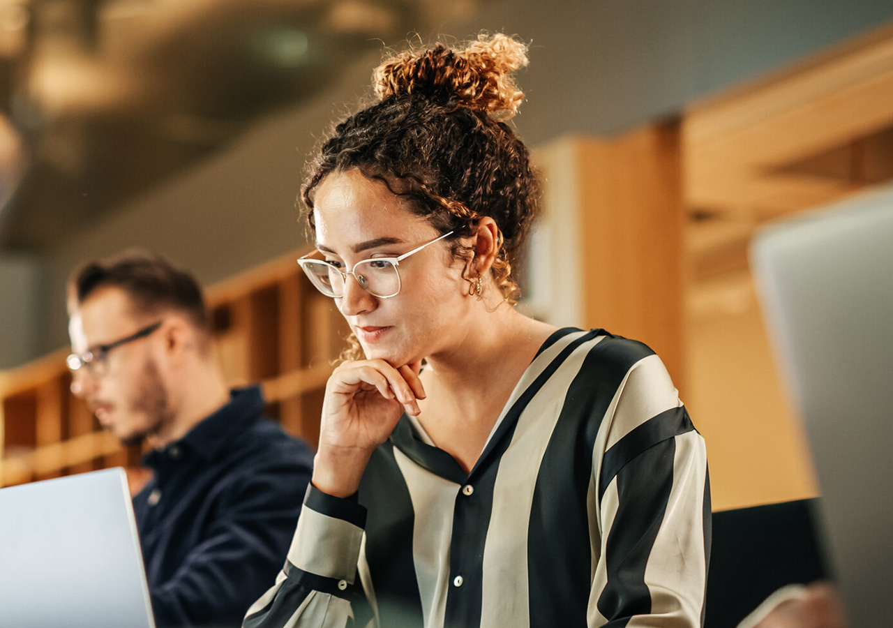 Eine Frau mit lockigem Haar und Brille, die eine schwarz-weiß gestreifte Bluse trägt, sitzt an einem Schreibtisch und arbeitet an ihrem Laptop - vielleicht aktualisiert sie einen Blog - in einem hellen, modernen Büro. Eine weitere Person arbeitet im Hintergrund.