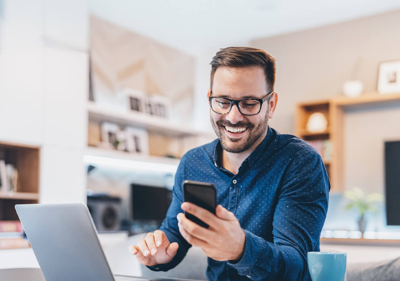 Ein lächelnder Mann mit Brille sitzt an einem Schreibtisch und nutzt ein Smartphone mit eCall, einen geöffneten Laptop und eine blaue Kaffeetasse vor sich in einer modernen Homeoffice-Umgebung – ideal, um flexible Businesss Optionen zu erkunden.