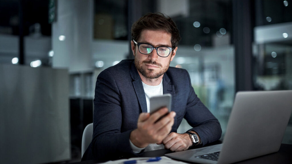 Ein Mann mit Brille und Blazer sitzt an einem Schreibtisch mit einem Laptop und schaut auf sein Smartphone. Das moderne Büro mit Glaswänden lässt vermuten, dass er Aufgaben für eCall - le service SMS suisse pour votre entreprise - verwaltet. Eine sanfte Beleuchtung vervollständigt die Szene.