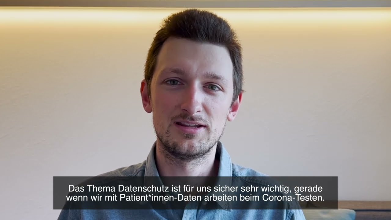 Ein Mann mit kurzen braunen Haaren und gestutztem Bart spricht vor einer beigen Wand mit einem horizontalen Lichtstreifen. Es werden deutsche Untertitel über die Bedeutung des Datenschutzes, insbesondere beim Umgang mit Patientendaten bei Corona-Tests, eingeblendet.